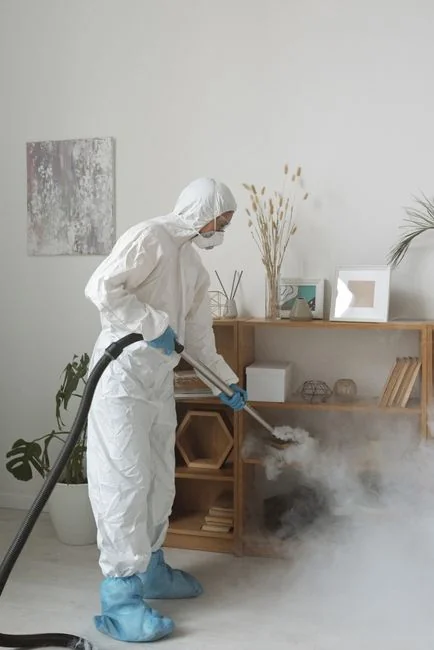 A person in protective gear sanitizing an office desk using fumigation equipment during daytime.